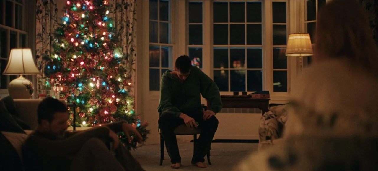 Babygirl (2024) movie still: medium — A dimly lit room at night features a large Christmas tree decorated with multicolored lights. Three people sit…