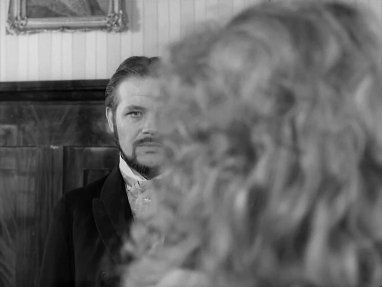 Effi Briest (1974) movie still: medium — A dimly lit room features a man focused on a person in the foreground. Blonde hair fills the right side of…