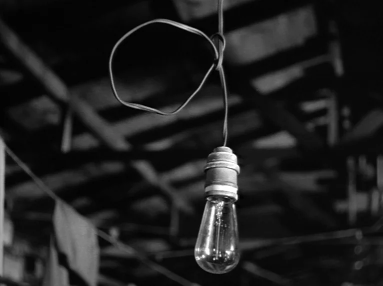Stalag 17 (1953) movie still: closeup — A bare incandescent bulb hangs from a twisted wire against a dark wooden ceiling. The rafters form a…
