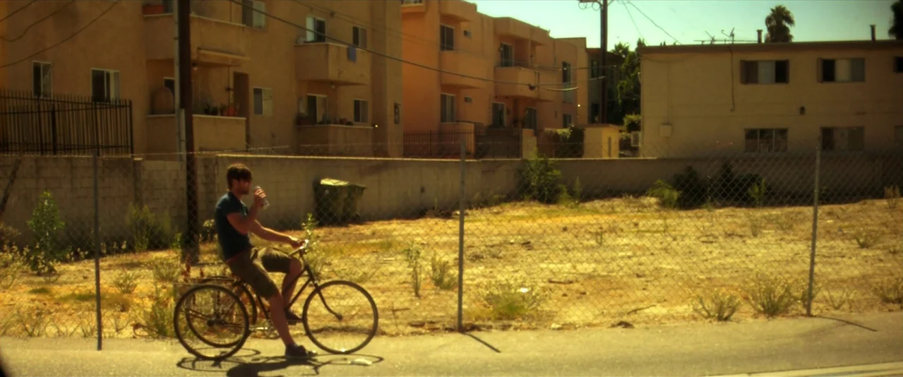 Bellflower (2011) movie still: wide — A sunny, dry urban vacant lot with a chain-link fence in the foreground. A man on a bicycle rides past the lot…