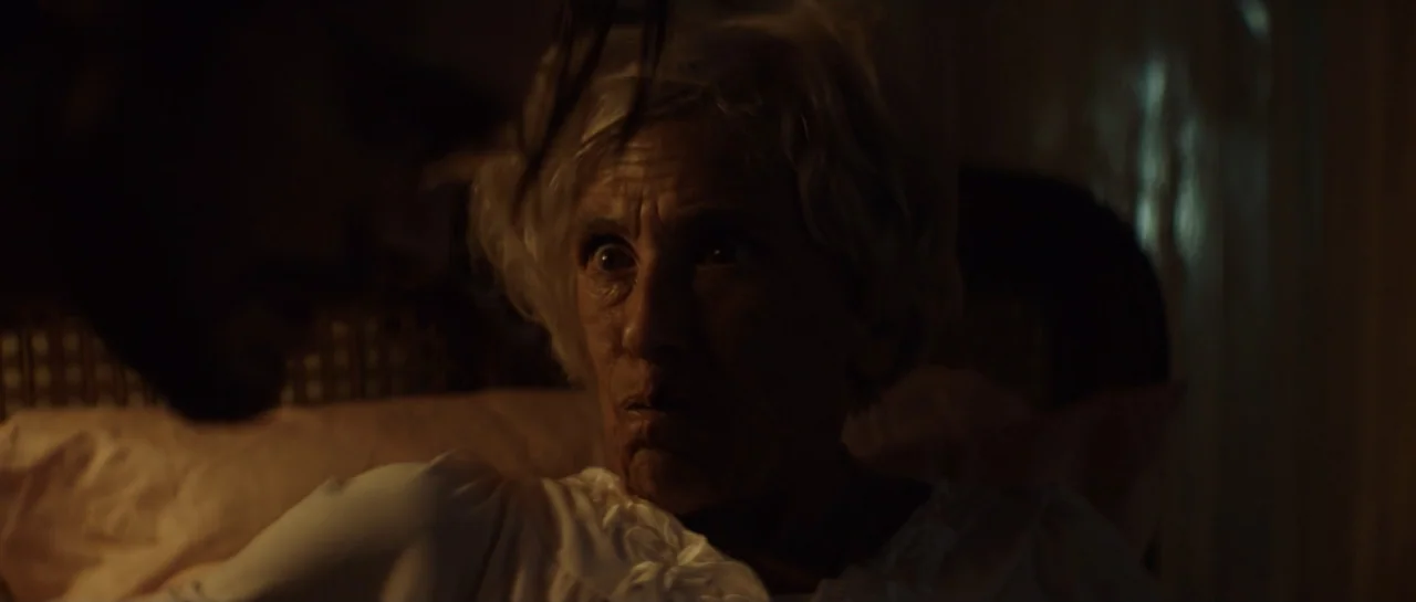The Front Room (2024) movie still: closeup — An interior bedroom at night with dark shadows and a dimly lit bed area. An elderly woman lies in bed…