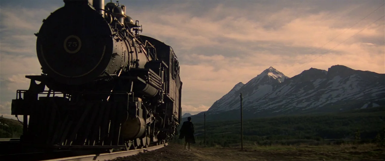 Heaven’s Gate (1980) movie still: wide — A steam locomotive sits on tracks against a large snow-capped mountain range at sunset. A lone figure walks…