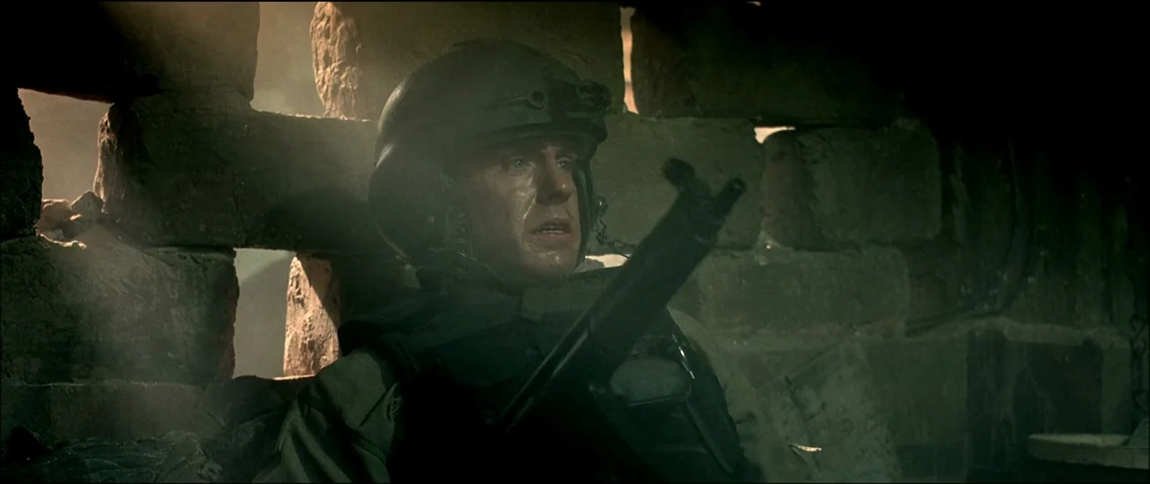 Black Hawk Down (2001) movie still: medium — A soldier is positioned within a gap of a crumbling brick structure during the day. He holds an assault…