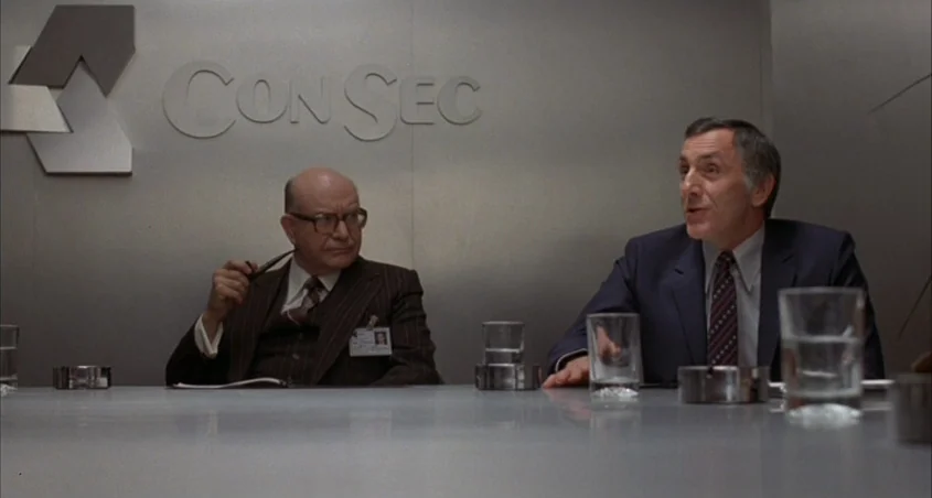 Scanners (1981) movie still: medium — A corporate conference room features a silver wall with a large ConSec logo signage. Two older men sit at a…