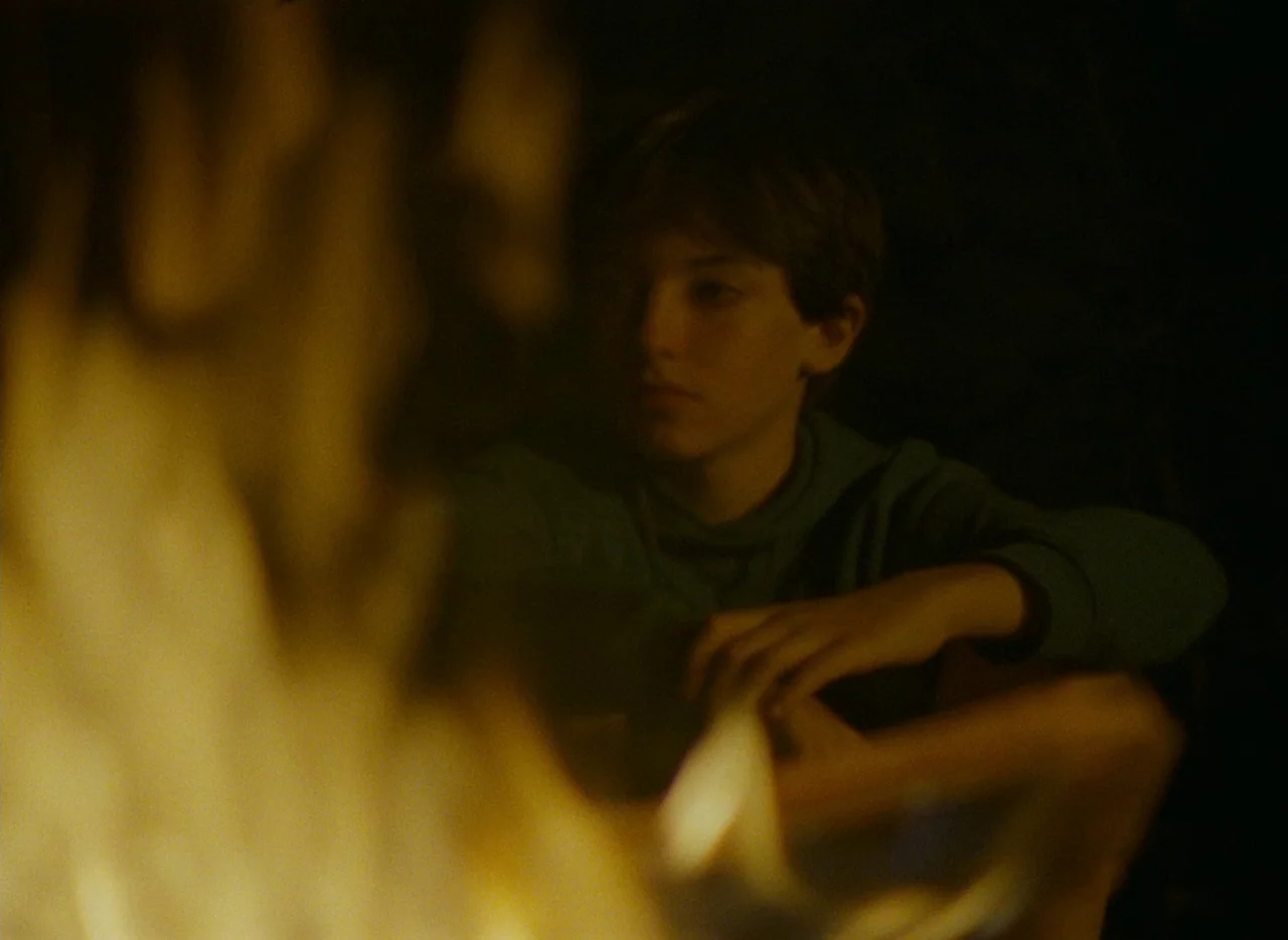 Falcon Lake (2022) movie still: medium — An outdoor area at night is illuminated primarily by a bright, blurry campfire foreground. A young boy in a…