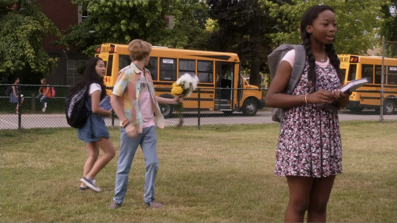 Rufus 2 (2017) movie still: medium — A grassy school lawn features a yellow bus parked behind a chain-link fence. Three students stand in the…