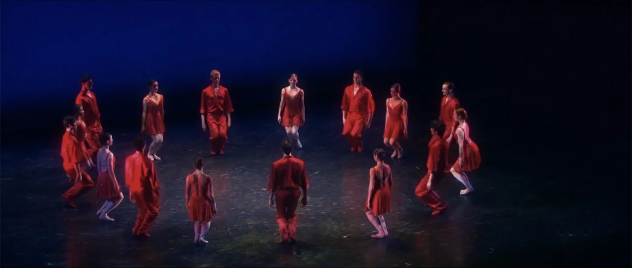 El acto (1989) movie still: wide — A dark, bare performance space holds a large group of dancers in red clothing. The performers are arranged in a…