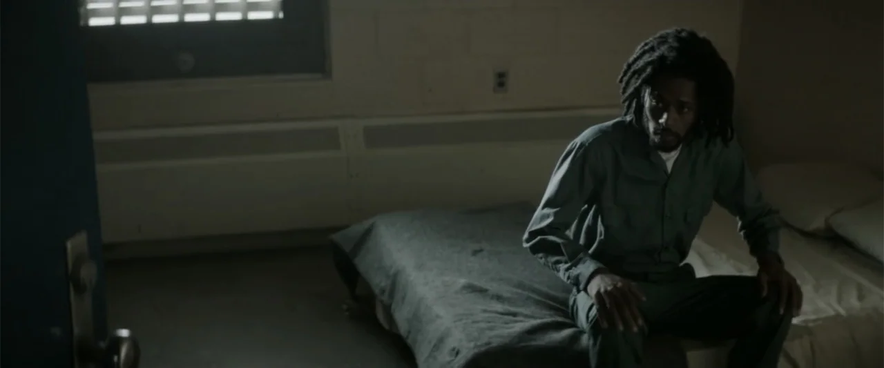 Crown Heights (2017) movie still: medium — A austere prison cell features a narrow bed against a beige painted wall. A man sits on the edge of the…