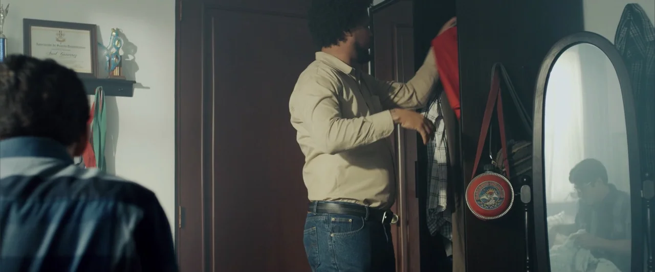 La Musiquita por Dentro (2019) movie still: medium — A bedroom contains an open wardrobe and a standing oval mirror near the wall. A man searches…