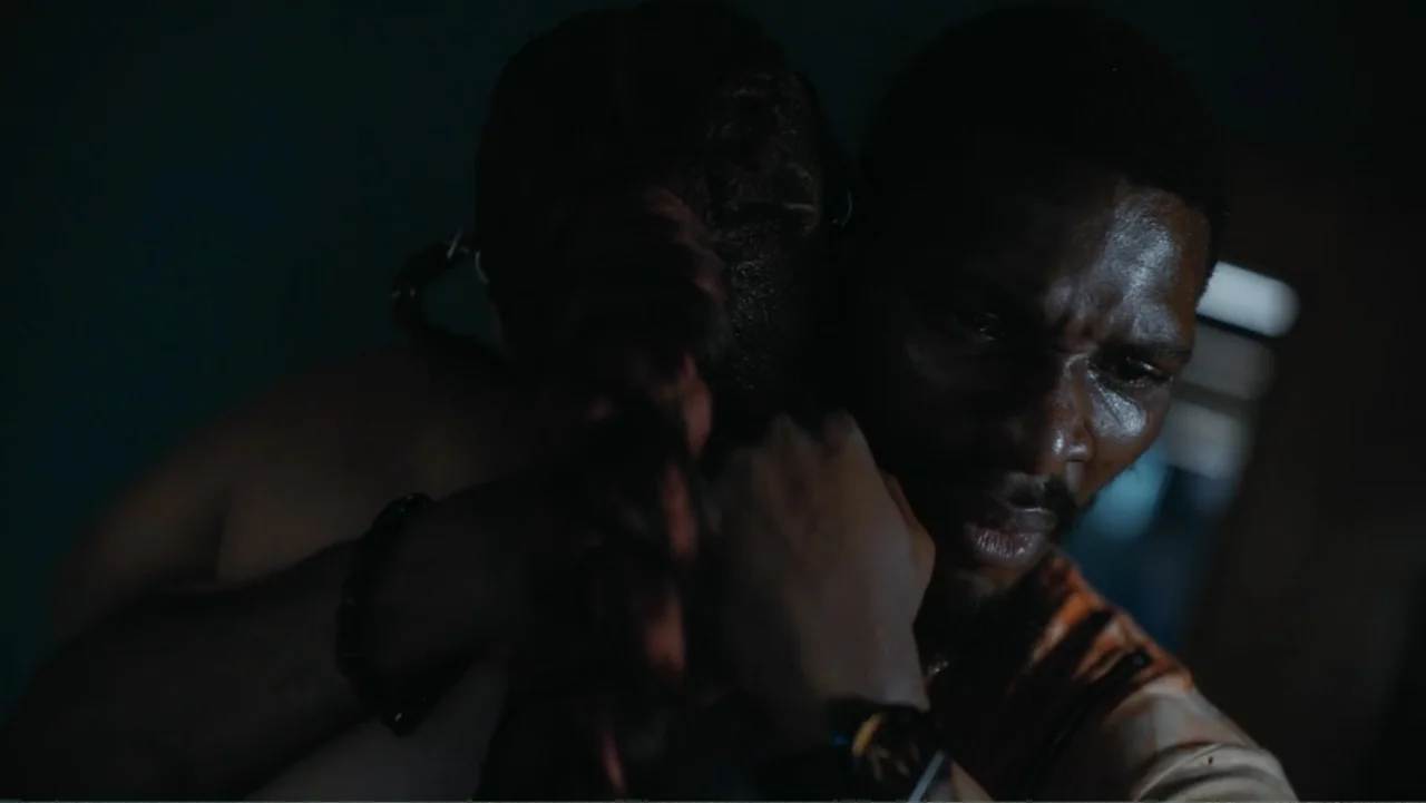 Gangs of Lagos (2023) movie still: closeup — A dark, dimly lit interior room with a single window source providing illumination. A man holds another…
