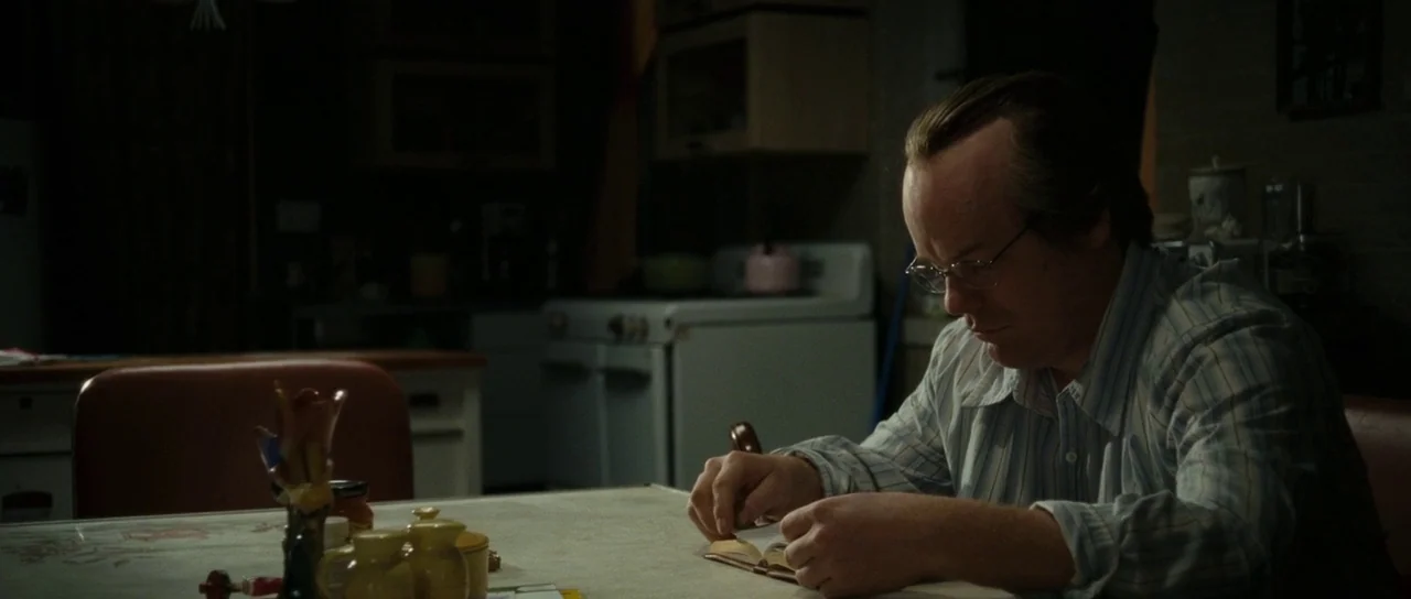 Synecdoche, New York (2008) movie still: medium — A man sits at a worn table in a dimly lit, dated room. He focuses intently on a small book while…