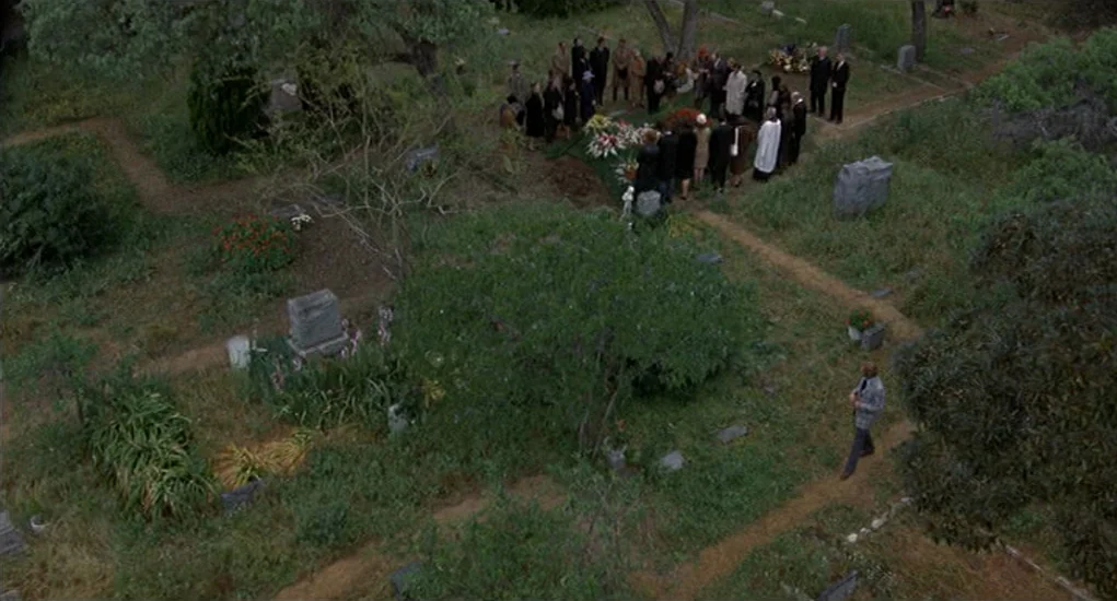 Family Plot (1976) movie still: wide — A group of mourners stands clustered around an open grave in a grassy cemetery. A single man walks along a…