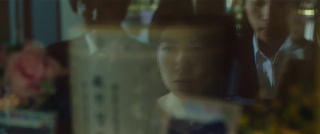 Psychokinesis (2018) movie still: closeup — A glass pane reflects a person gazing at an arranged memorial with funeral offerings. The subject's face…