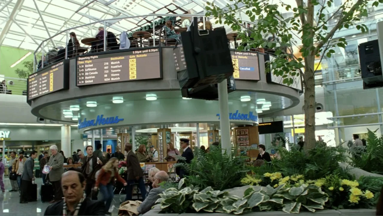 The Terminal (2004) movie still: wide — A busy airport terminal interior featuring a circular newsstand and digital flight departure monitors.…