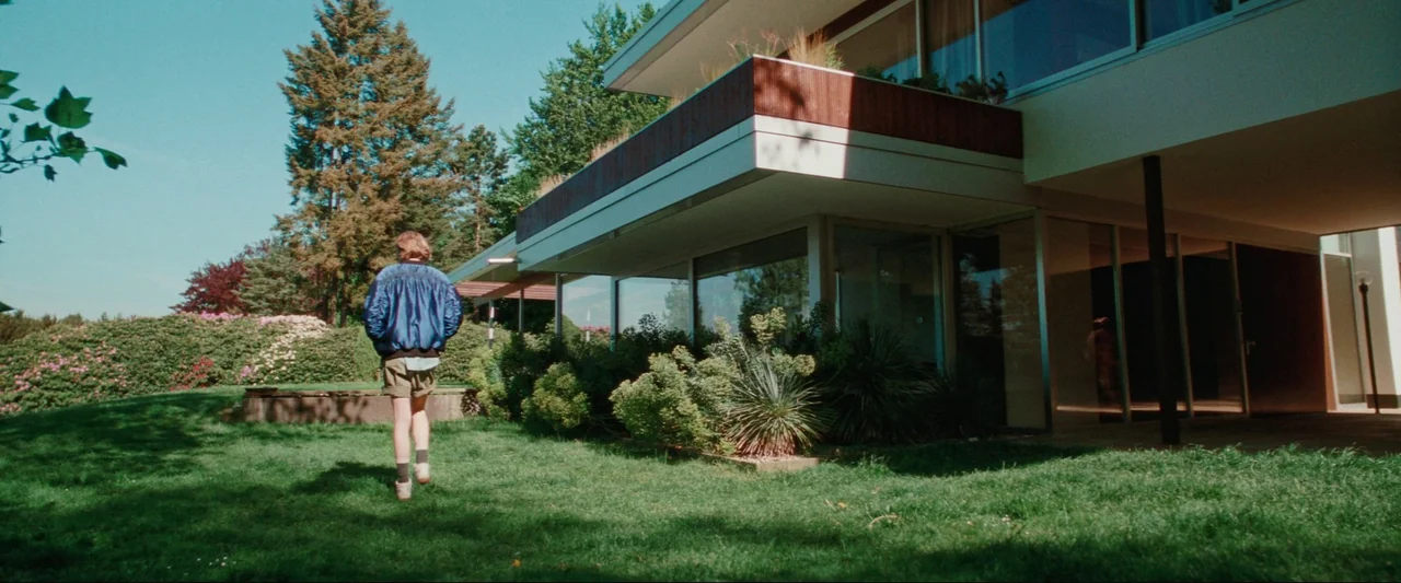 Cuckoo (2024) movie still: fullbody — A modernist house with large glass windows stands beside a manicured green lawn. A person in a blue jacket…