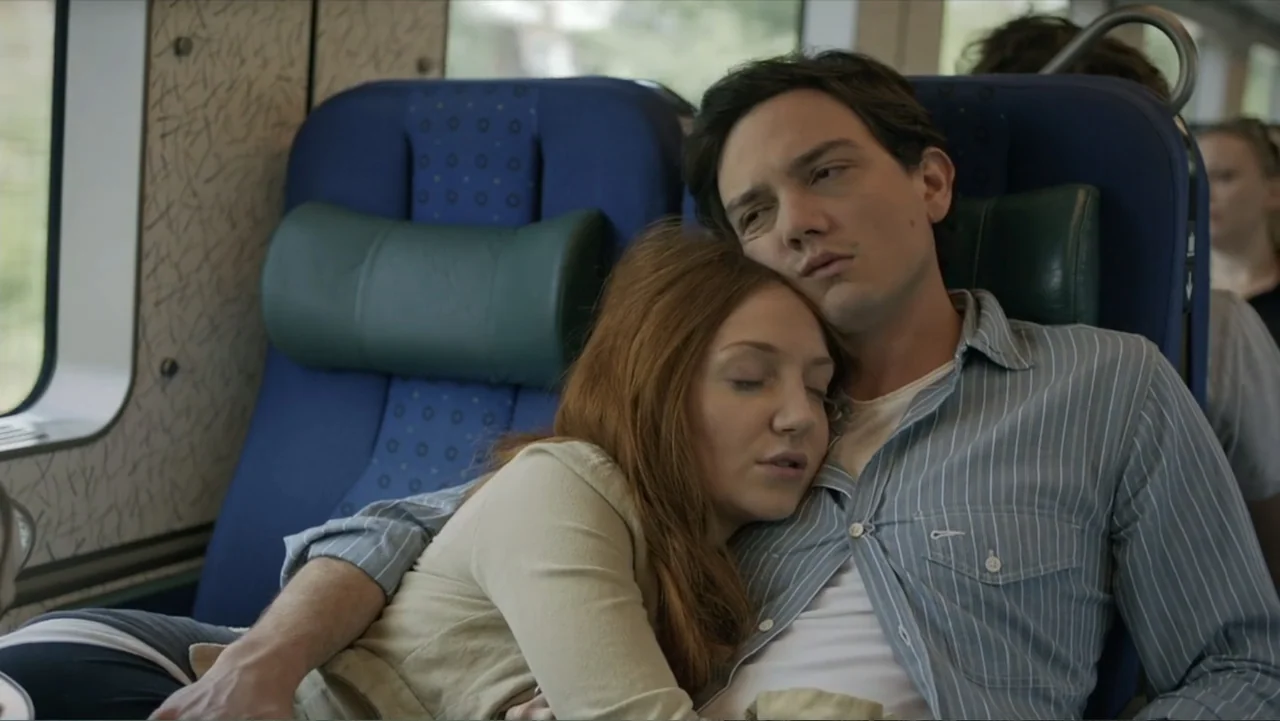 Copenhagen (2002) movie still: medium — The interior of a passenger train car features blue upholstered seats with headrests. A young man and woman…