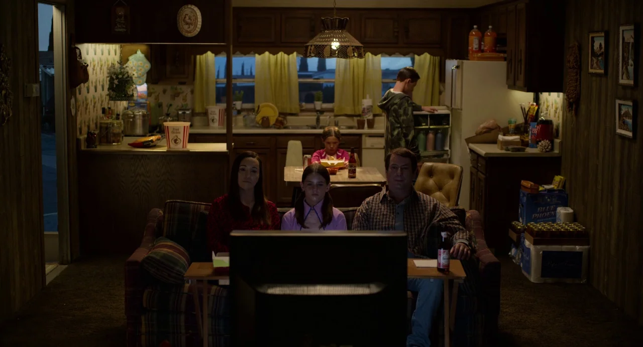 Velvet Buzzsaw (2019) movie still: wide — A 1990s mobile home living room featuring wood-paneled walls and dated laminate cabinetry. Three family…
