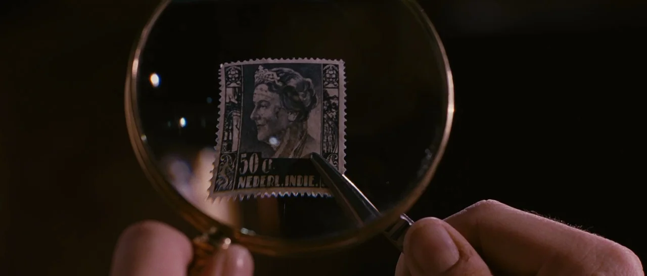 Black Book (2006) movie still: closeup — A vintage postage stamp from the Dutch East Indies sits inside a magnifying glass. A hand holds the…