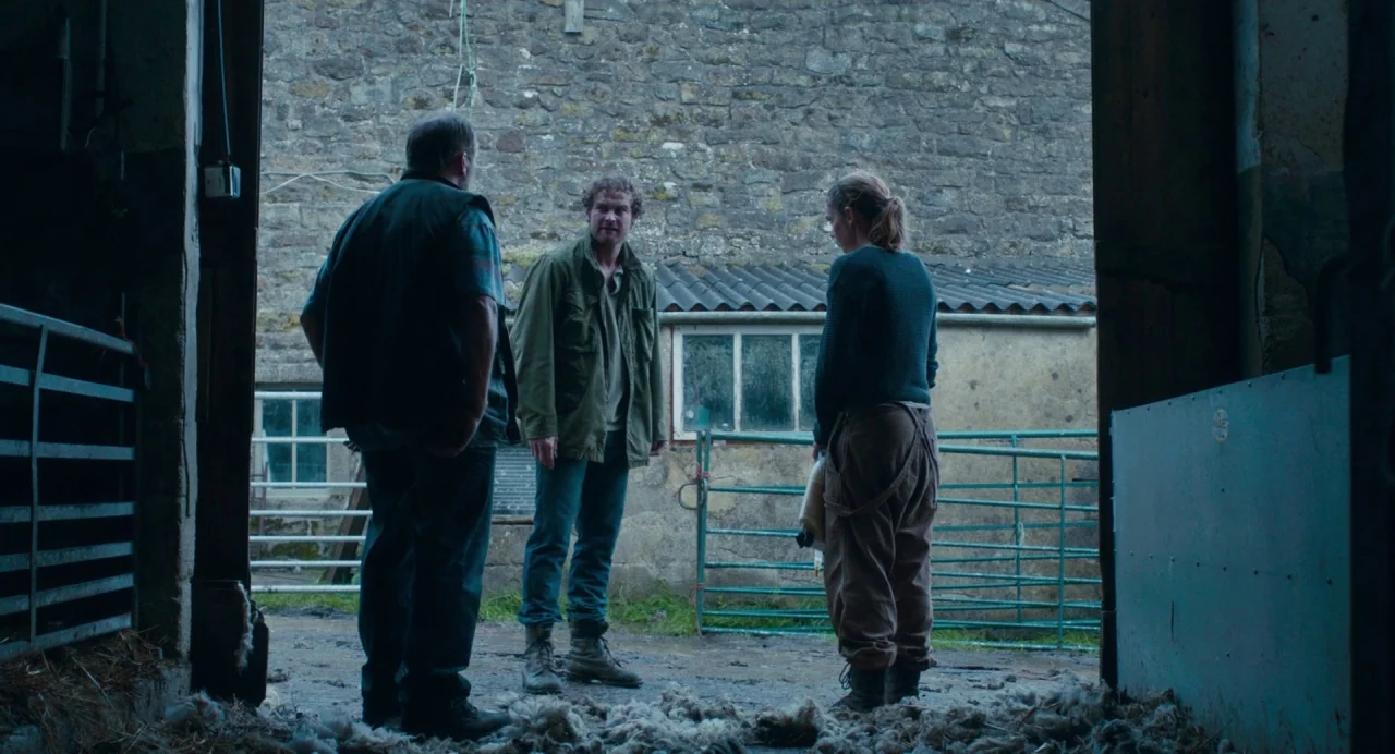 Dark River (2017) movie still: fullbody — A stone-walled agricultural structure with an open entrance faces a courtyard area. Three people converse…