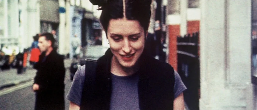 Wonderland (1999) movie still: medium — A woman walks down a city street lined with buildings and brick pillars. Another person stands blurred in…
