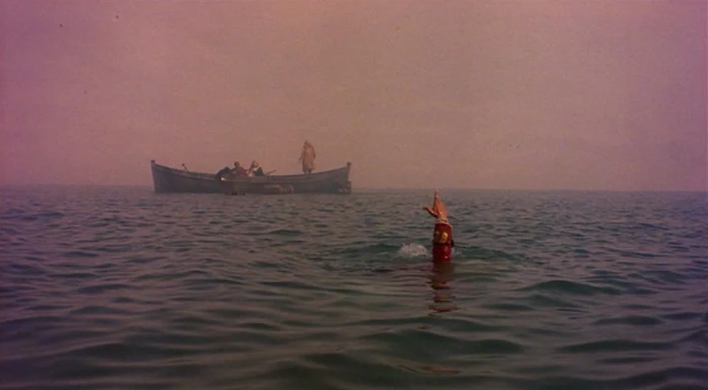 The Adventures Of Baron Munchausen (1988) movie still: wide — A hazy seascape features a wooden boat occupied by several figures in the distance. An…
