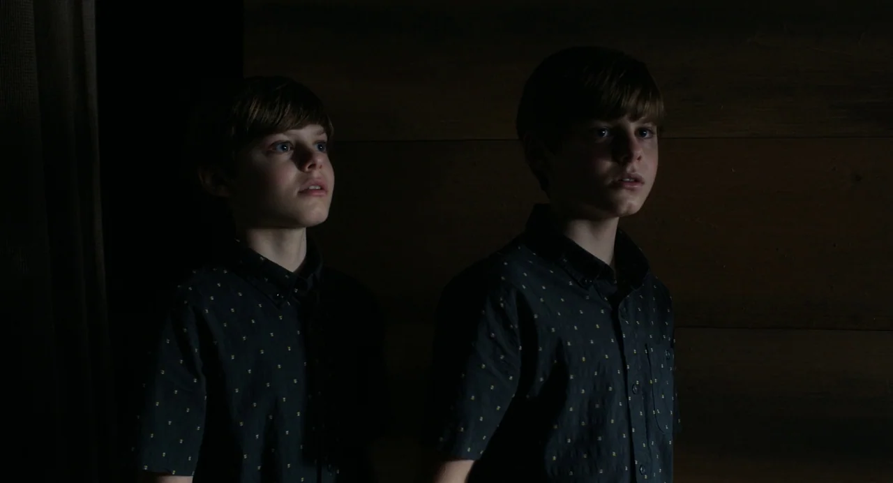 Goodnight Mommy (2022) movie still: medium — Two identical young boys stand against a dark wood-paneled wall in a dim space. They gaze toward the…