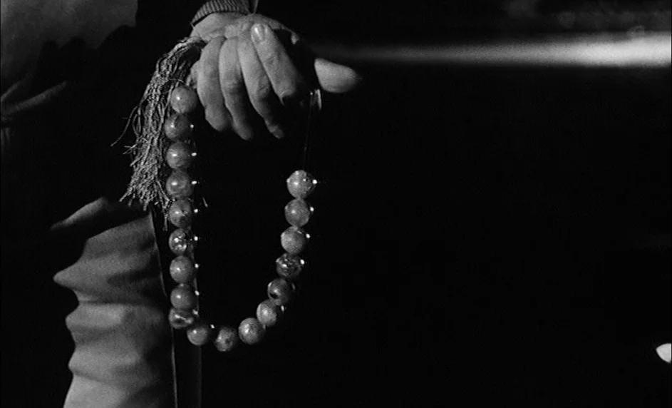 Boy Meets Girl (1984) movie still: closeup — A close-up shot of a hand holding a strand of prayer beads. The background is completely dark with no…