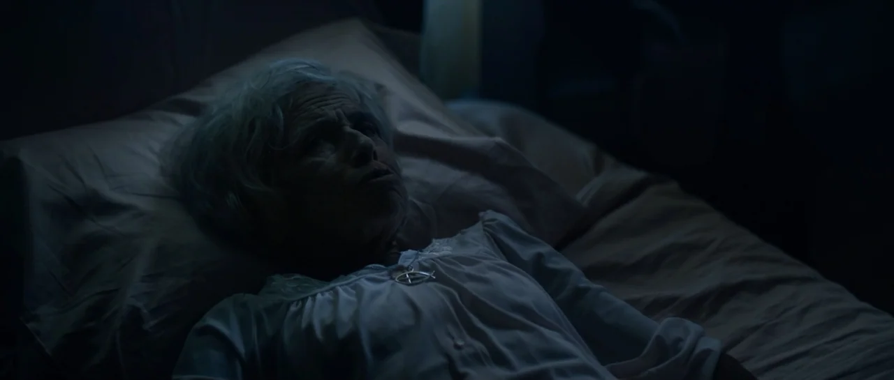 The Front Room (2024) movie still: closeup — An interior bedroom at night features an elderly woman lying on a bed. The scene is dominated by…
