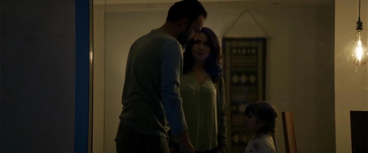 Human Animals (2020) movie still: medium — A dimly lit room interior features a decorative wall hanging and a hanging bulb. A man and woman stand…