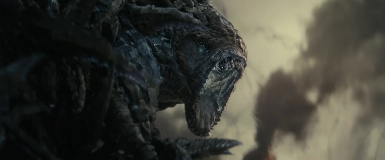 Edge of Tomorrow (2014) movie still: closeup — A jagged creature stands amid a dark, smoky environment with floating debris. The monster tilts its…