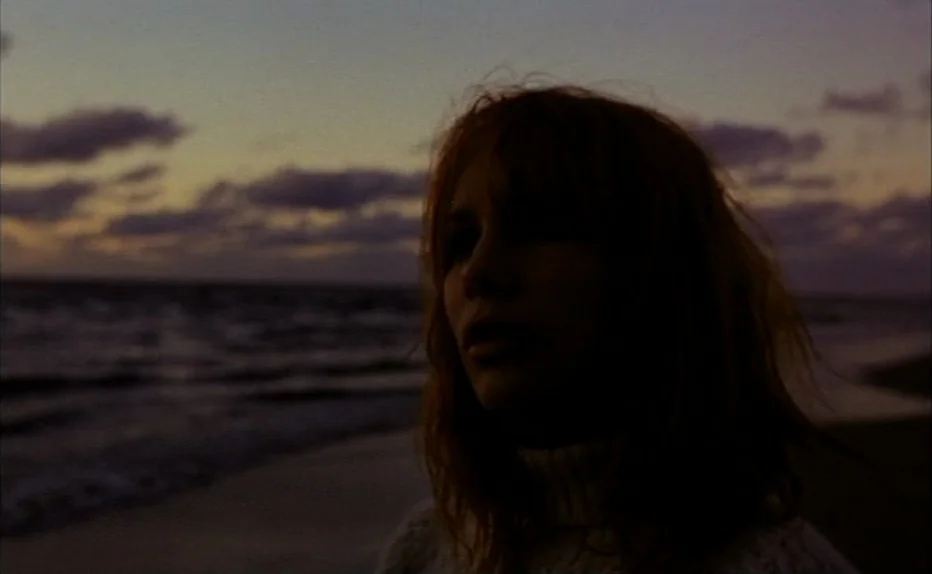 Turkish Delight (1973) movie still: closeup — A beach shoreline at dusk under a dark, cloudy sky. A young woman stands facing the horizon while the…
