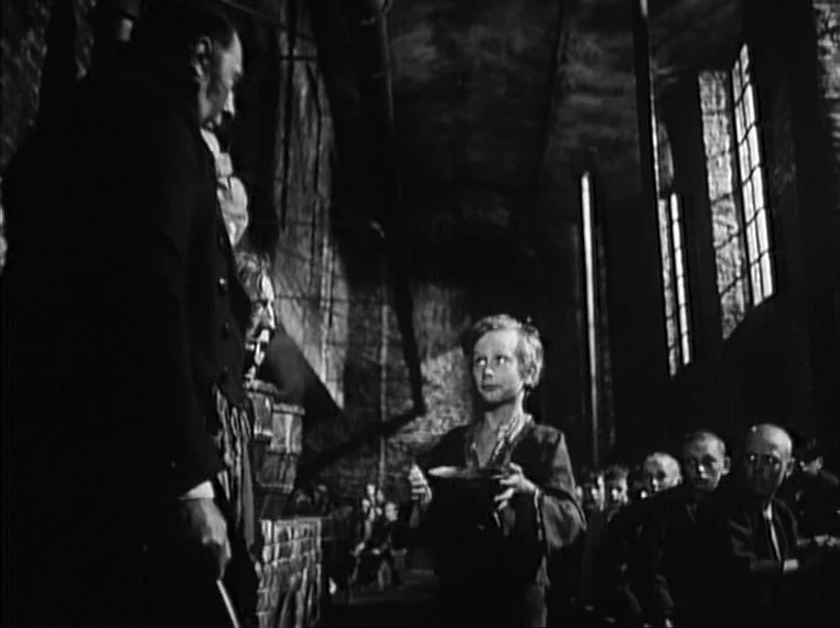 Oliver Twist (1948) movie still: medium — A dark stone hall with high windows and long rows of seated people. A young boy stands before a man…