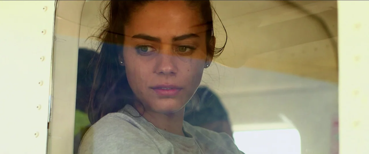 Green Inferno (2013) movie still: closeup — A woman looks out through the reflective glass of an airplane window. The interior cabin walls are…