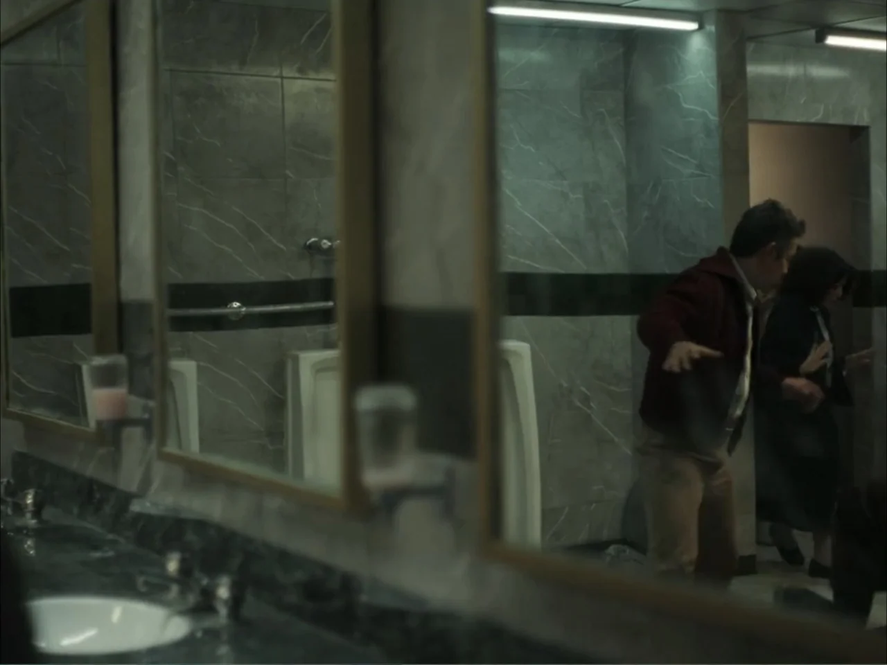 November (2025) movie still: medium — A public washroom features gray marbled wall tiles and a long mirror reflection. A man in a burgundy jacket…