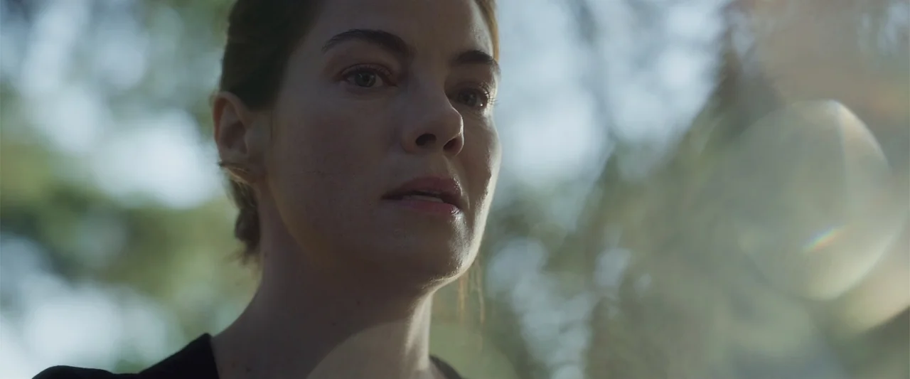 Black Site (2022) movie still: closeup — A woman stands in a blurred outdoor area with trees in the background. She looks toward the side of the…