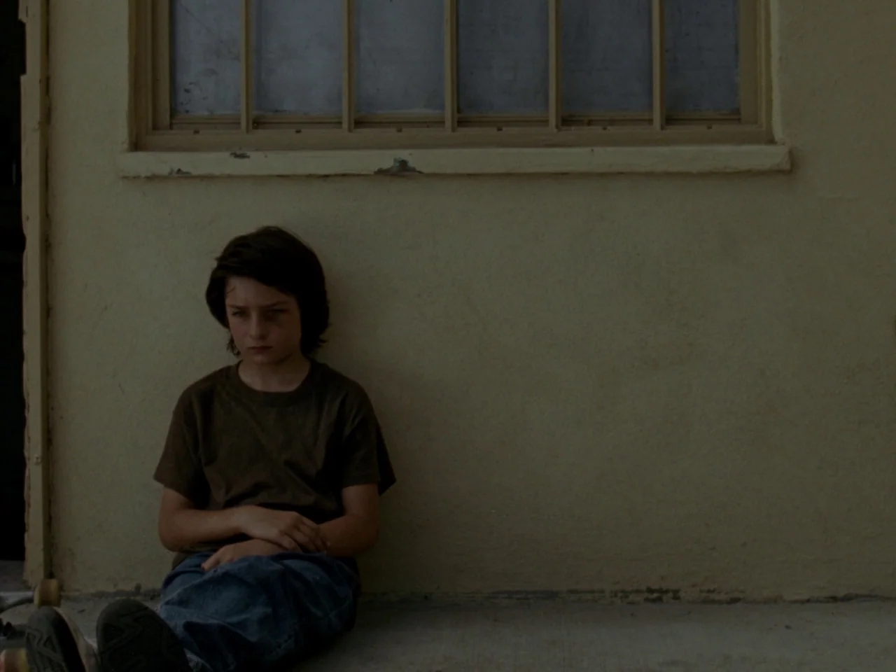 Mid90s (2018) movie still: medium — A boy sits on a concrete ground against a weathered yellow stucco wall. A small window with a wooden frame is…