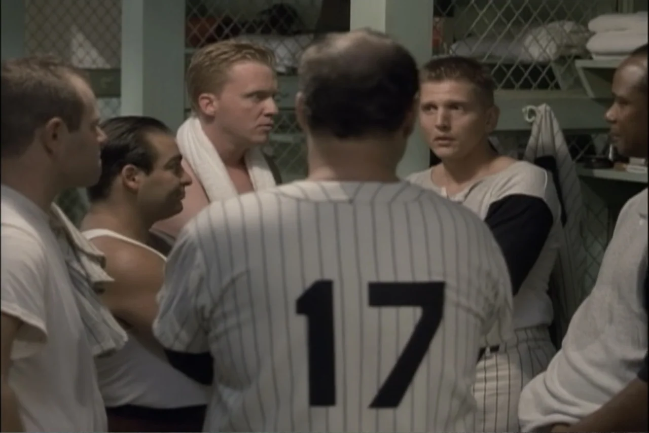 Unification of Japan 61 (2024) movie still: medium — A sports team gathers in a cramped interior locker room with wire mesh lockers. Several male…