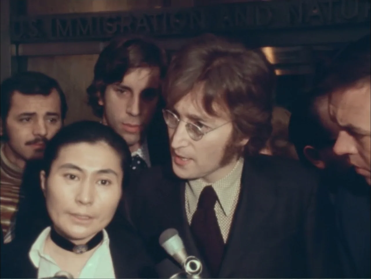 One to One: John & Yoko (2025) movie still: medium — A crowded hallway in an official building with dark wood panels and lettering visible. John…