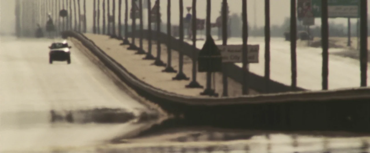 Syriana (2005) movie still: wide — A long, multi-lane desert highway stretches into the distance under a thick, hazy sky. A single dark car travels…