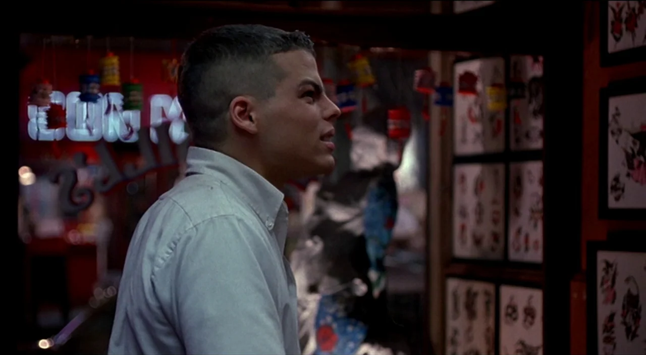 Dogfight (1991) movie still: medium — A dimly lit room displays various flash art prints in black frames on red walls. A young man with a…
