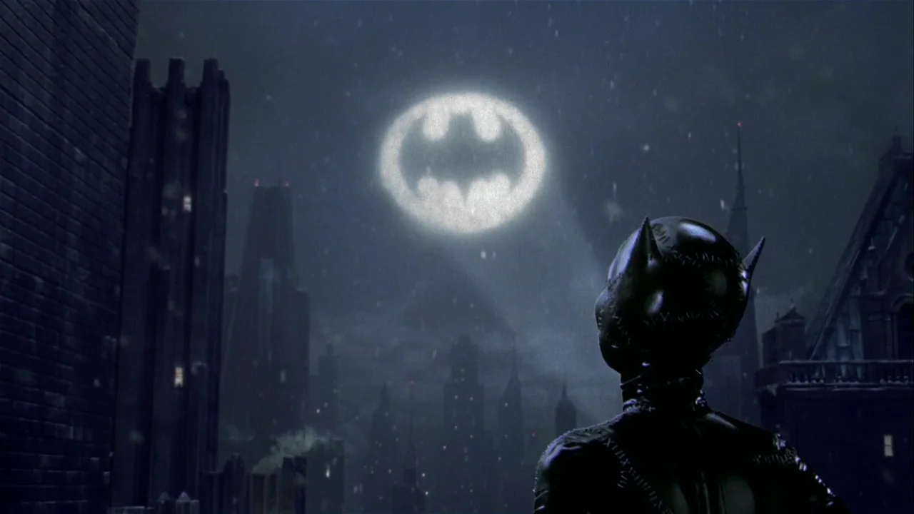 Batman Returns (1992) movie still: medium — A dark, snowy urban rooftop overlook at night features gothic architecture and city buildings. A figure…