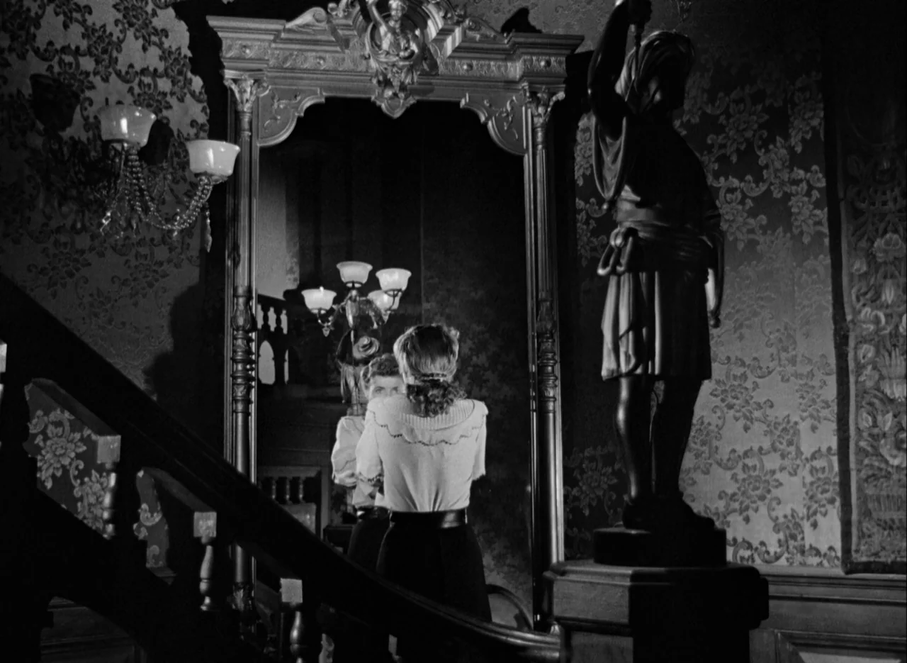 The Spiral Staircase (1946) movie still: medium — A dimly lit grand staircase features floral wallpaper, a tall sculpture, and a large mirror. A…