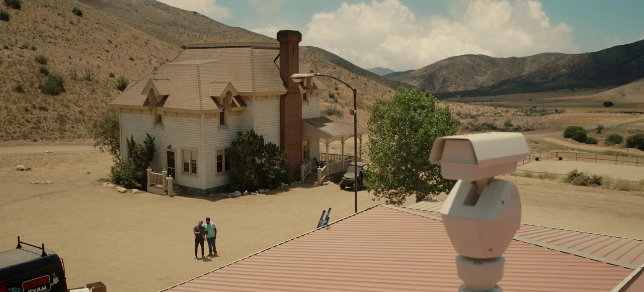 Nope (2022) movie still: wide — A rural ranch estate sits in a dry, mountainous desert landscape under clear skies. Two men converse in the dirt…