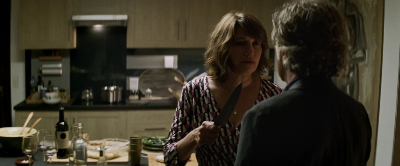 Masquerade (2022) movie still: medium — A modern domestic cooking space features wood-toned cabinetry and a countertop with food items. A woman in a…