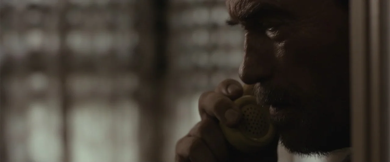Maggie (2015) movie still: closeup — A dimly lit room features a subject framed against a blurred window background. A man holds a vintage telephone…