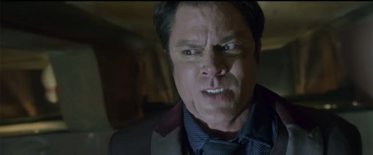 Skiptrace (2016) movie still: closeup — A dimly lit room interior with dark shadows obscuring the background. A man in his forties looks off-camera…