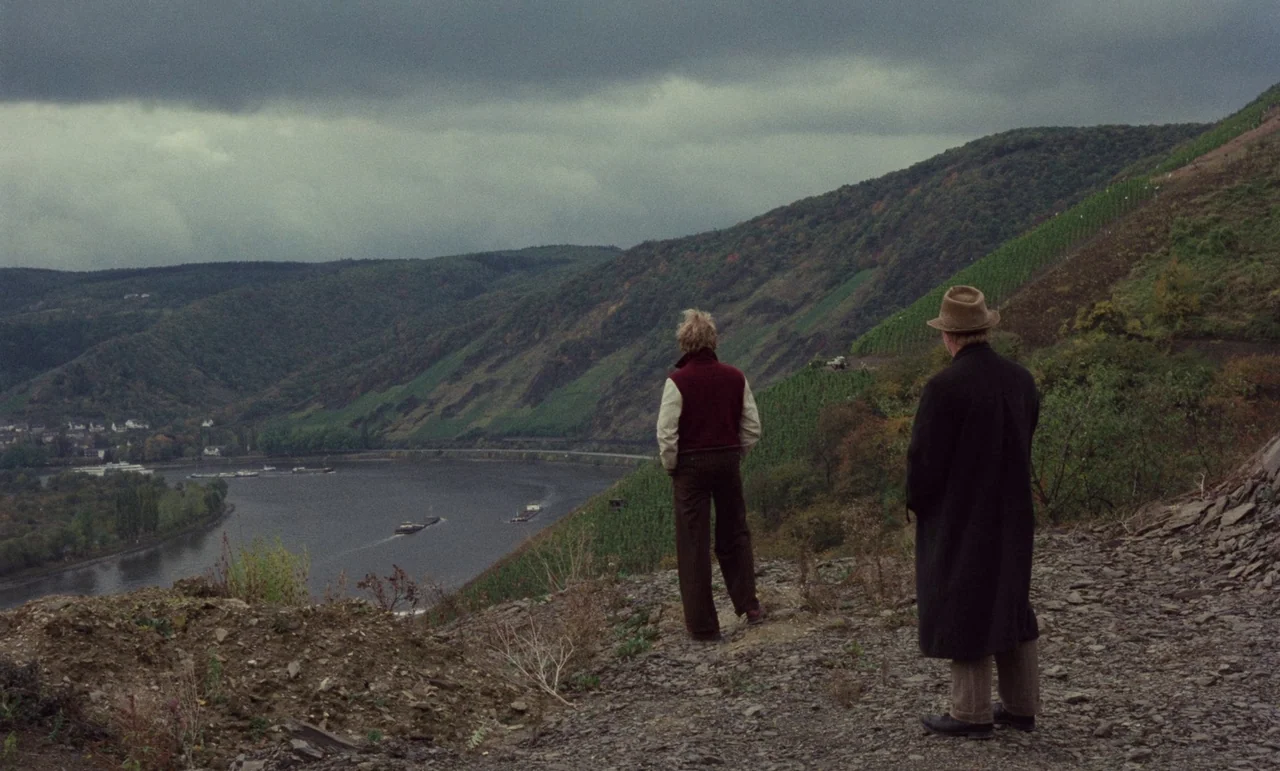Wrong Move (1975) movie still: wide — A rocky viewpoint overlooks a wide river flowing through a hilly green valley. Two men stand together on the…