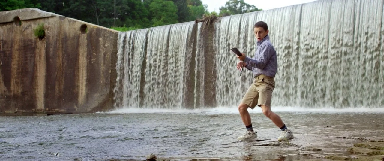 Kings Of Summer (2013) movie still: fullbody — A man-made concrete spillway creates a wide, flat waterfall into a shallow rocky river. A young man…
