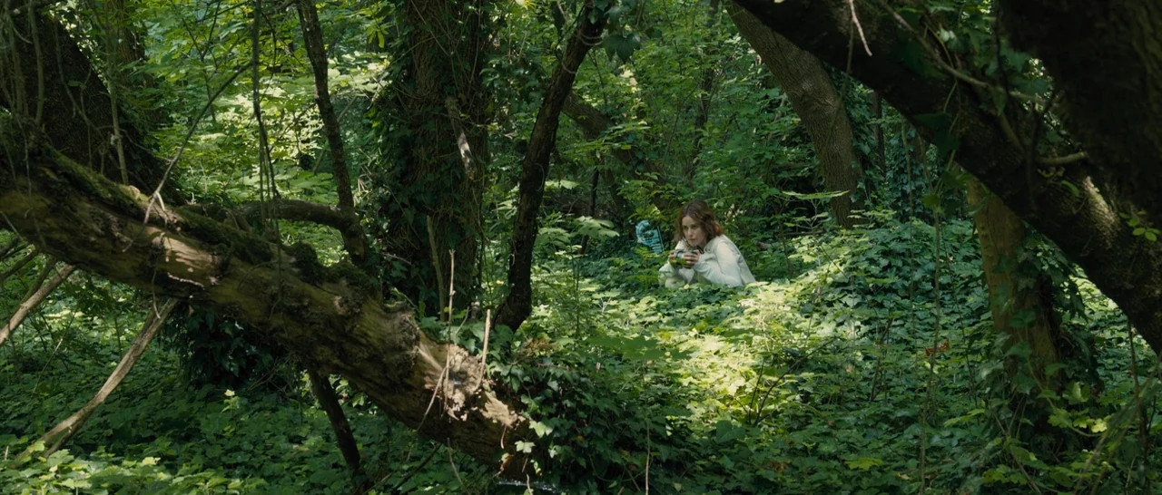 Blade Runner 2049 (2017) movie still: wide — A dense, overgrown wooded area features thick tree trunks and a carpet of ivy. A woman in a white…