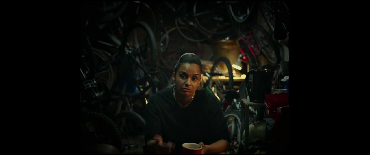 Scrapper (2023) movie still: medium — A dark, cramped room is filled with numerous bicycles hanging on the walls. A person sits in the foreground…